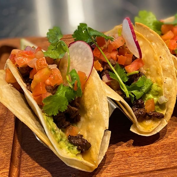 Two Culichis Urban Kitchen tacos with meat, guacamole, pico de gallo, cilantro, and radish on a wooden board.